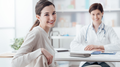 Eine Ärztin am Schreibtisch lacht mit ihrer Patientin in die Kamera